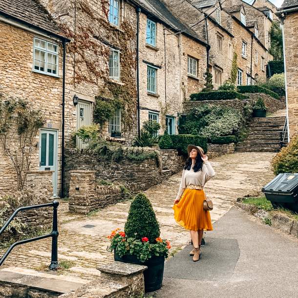 A woman wearing a hat smiles as she poses in front of stone cottages on a stepped hill in Tetbury