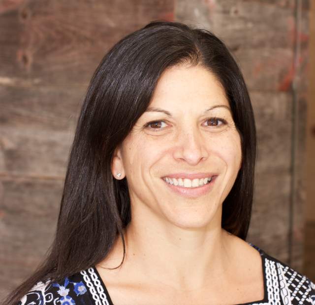 Headshot photo of a smiling woman with dark brown hair and brown eyes. She is wearing a blue and white patterned blouse with a squared neckline