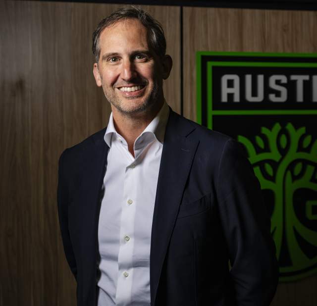 Photo of a man in a black blazer and white undershirt smiling into the camera posing for a headshot with a large Austin FC logo on the wall behind him.
