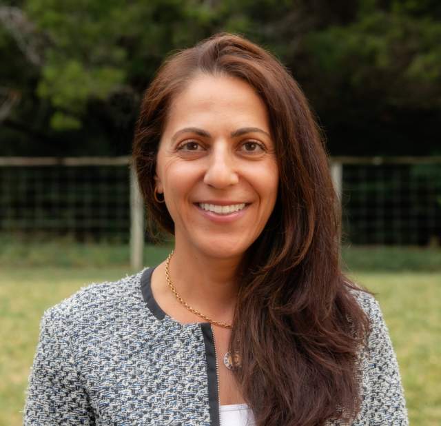 Photo of a woman with dark brown hair standing outside wearing a blazer standing and smiling into the camera posing for a headshot.