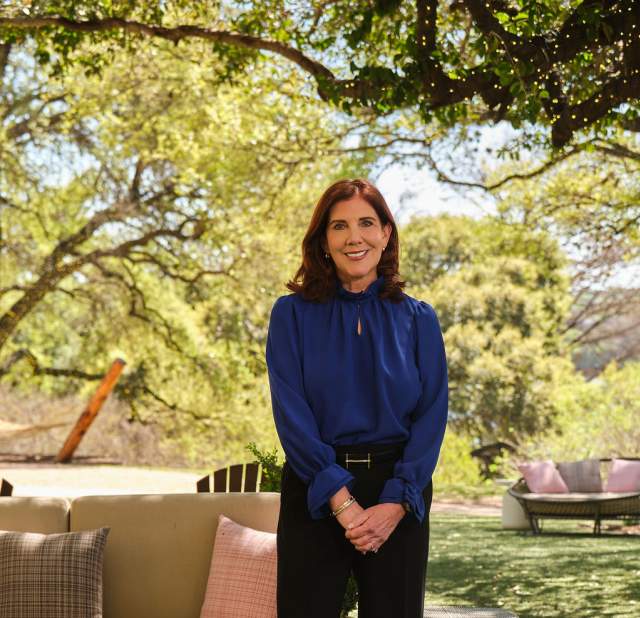 A woman wearing a blue top and black slacks in an outdoor setting smiling into the camera posing for a headshot.