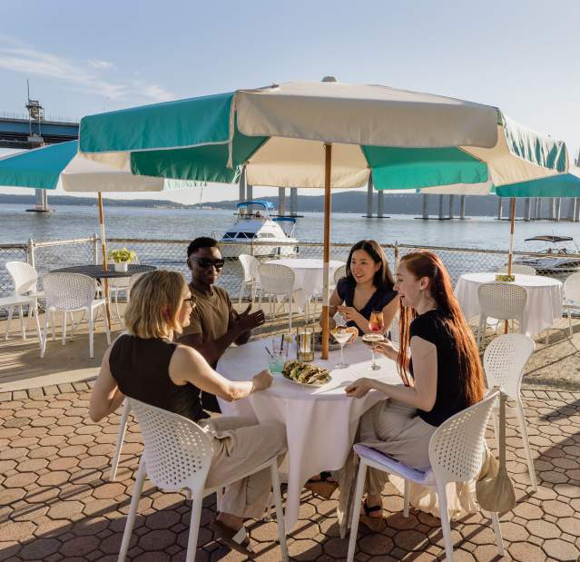 A group of four people sit at an outdoor dining table overlooking the Hudson River. They are laughing and chatting.
