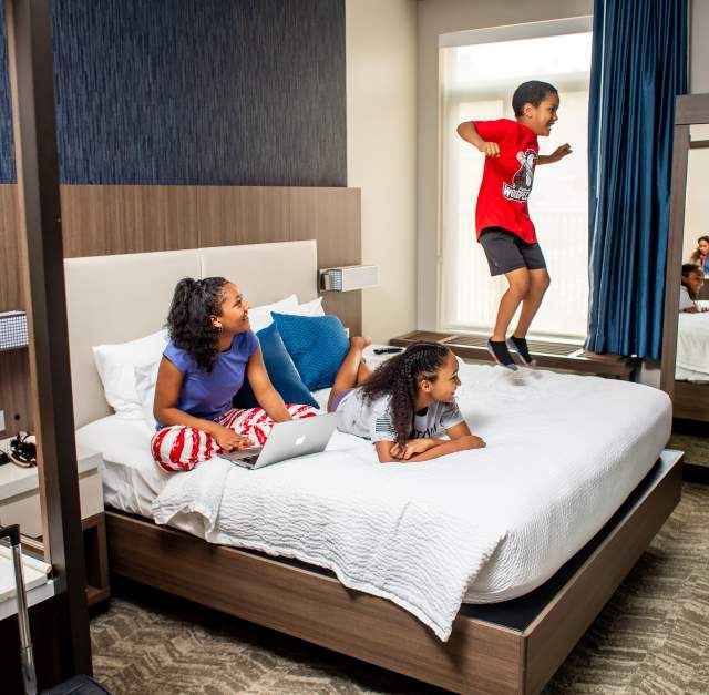 Family relaxing and playing inside a modern hotel room, with children jumping on the bed and parents enjoying a comfortable overnight stay.
