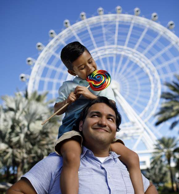 A boy on top of his dad's shoulders and eating a lollipop in front of The Wheel at ICON Park