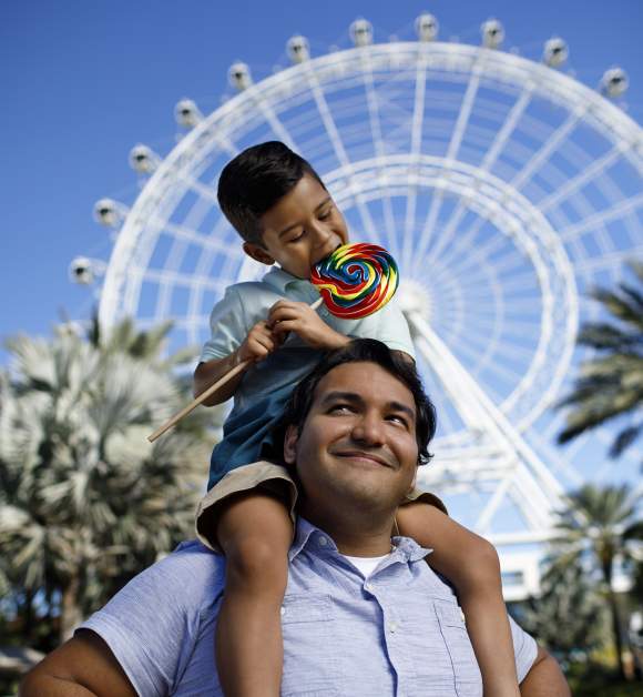 A boy on top of his dad's shoulders and eating a lollipop in front of The Wheel at ICON Park