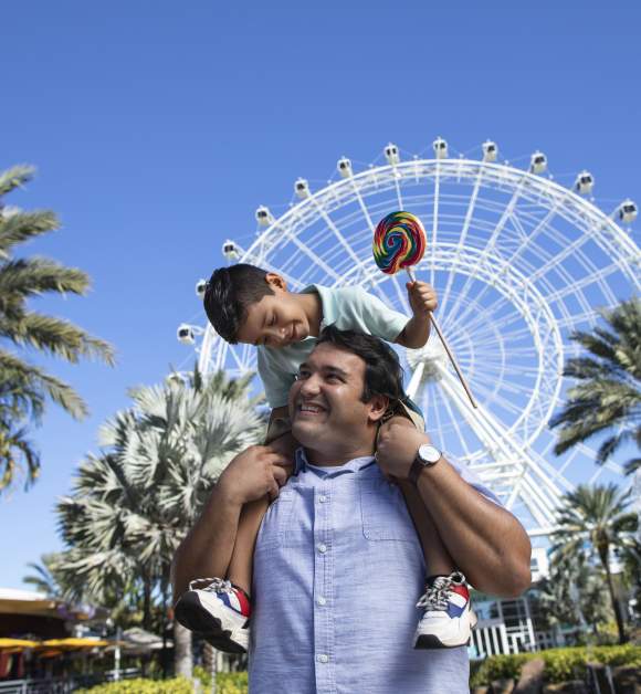 A boy on top of his dad's shoulders, while smiling and holding a lollipop, in front of The Wheel at ICON Park
