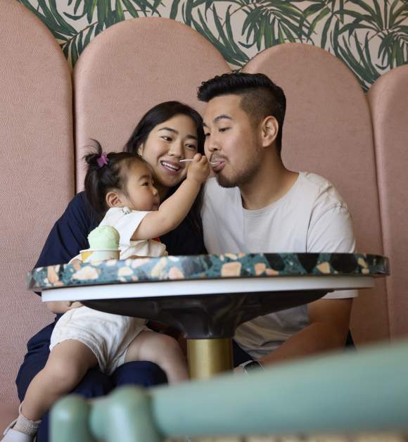 A family sitting together, with a toddler feeding the father in a cozy restaurant setting with pink seating and leafy wallpaper.