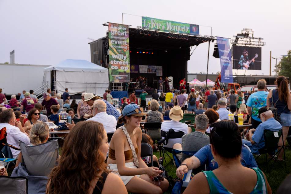 A large crowd sits and stands on a grassy area facing an outdoor concert stage at La Fête de Marquette, with musicians performing under stage lights and festival banners visible above the stage.