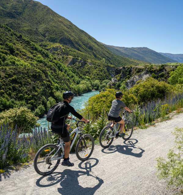 Two people biking along a blue river with wild flowers