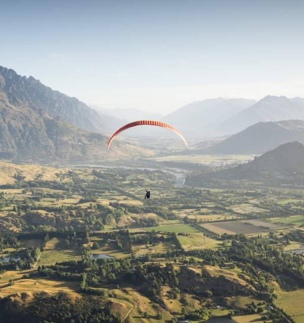 Person paragliding over a green valley with the Remarkables mountain range in the background