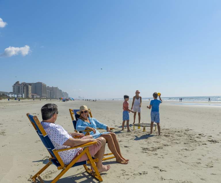 A mom and dad relax on the beach while their children play a beach game in the background.