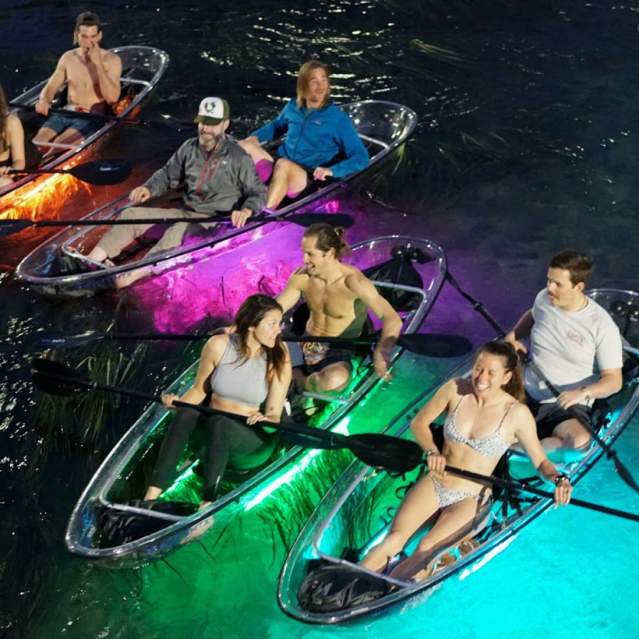 People paddling in clear kayaks at night with colored lights