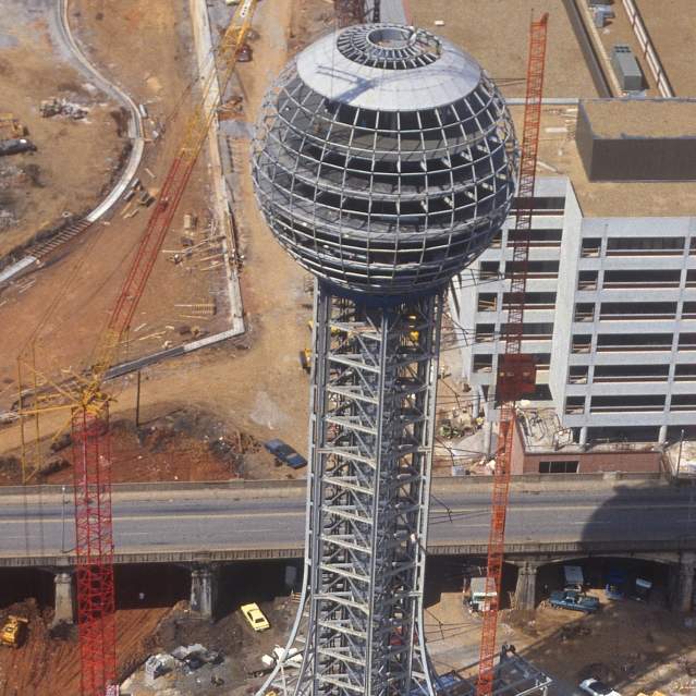 A high-angle historical photograph showing the Sunsphere under construction in Knoxville, Tennessee, for the 1982 World's Fair. The structural steel framework of the tower and the unfinished sphere are visible, surrounded by construction cranes, dirt lots, and neighboring buildings in progress.