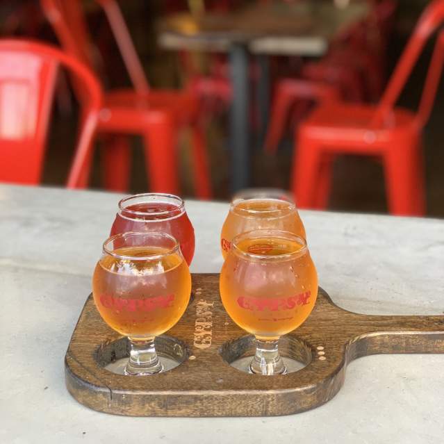 Four small glasses of craft beer arranged on a wooden tasting flight paddle on a light-colored table inside a softly lit bar. Bright red metal chairs and tables blur into the background, creating a warm, cozy atmosphere.