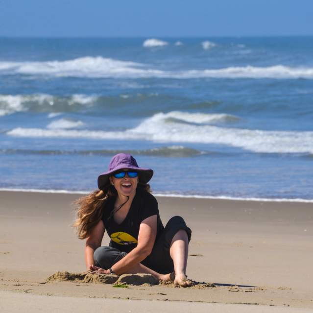 Woman sits building a sand castle on the beach with the ocean behind her. She is smiling under a large, purple brimmed hat.