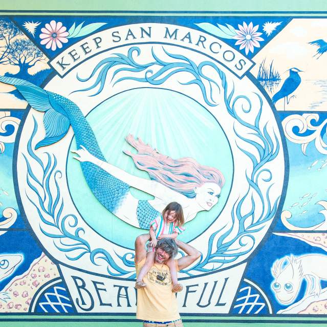 Father and daughter pose in front of a mural of a mermaid.