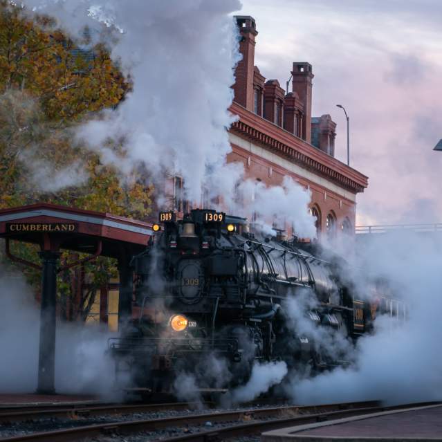 A large black steam train engine pulls into the Cumberland Station as it releases a billowy cloud of steam.