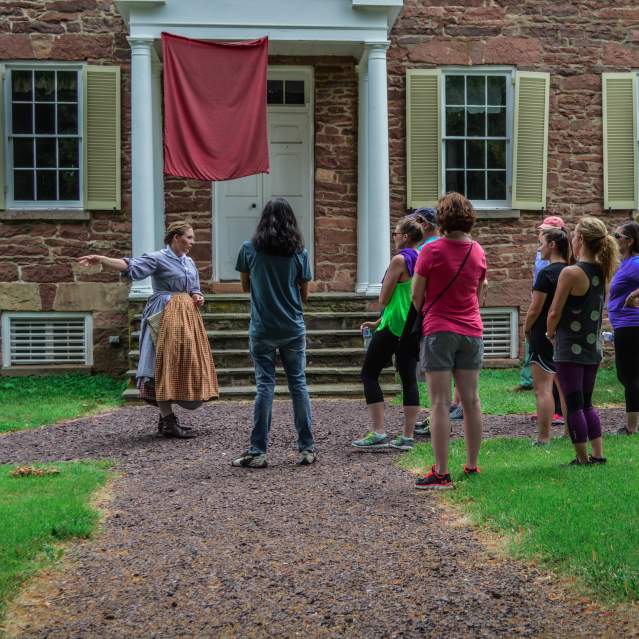 A group of visitors listening to a woman dressed in historic attire outside of a brick building