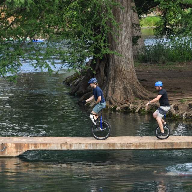 Unicycle riders on bridge over San Marcos River