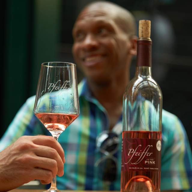 In the foreground there is a bottle and two glasses of rosé. The bottle says Pfeiffer. In the background and out of focus is a man smiling.