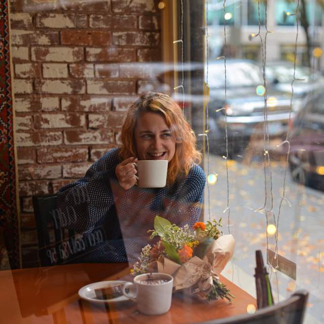 A person smiles with a cup of coffee near a coffee house window