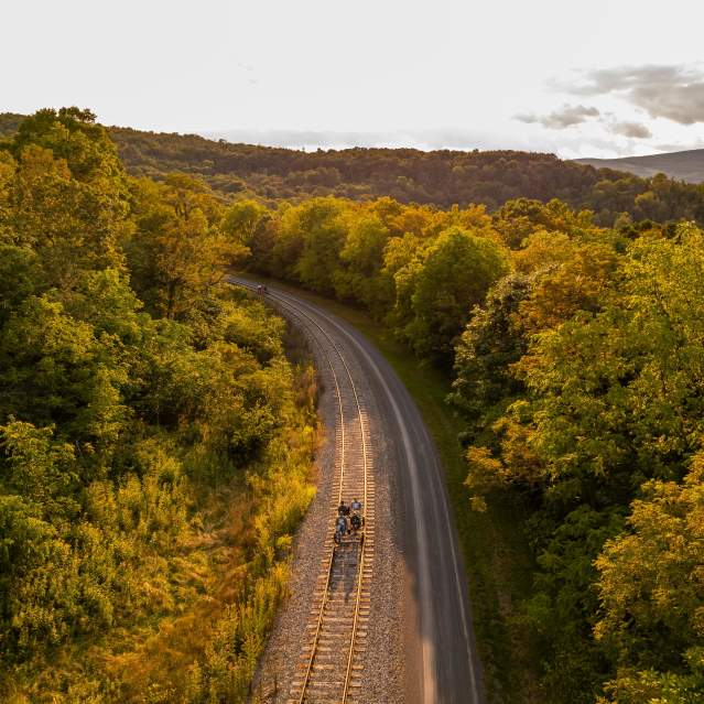 Four passengers peddle along a rail line on peddle bikes with the trees, sky and clouds in the background at sunset.