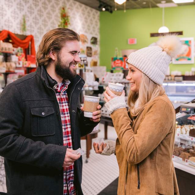 A smiling couple enjoys hot drinks inside a festive Kilwins decorated with holiday colors. They stand near a display case filled with assorted sweets and baked goods, creating a warm and cheerful atmosphere.