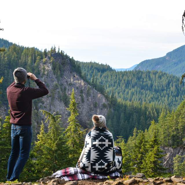 Two people sit on the edge of a forested cliff looking out into the Cascade Mountains