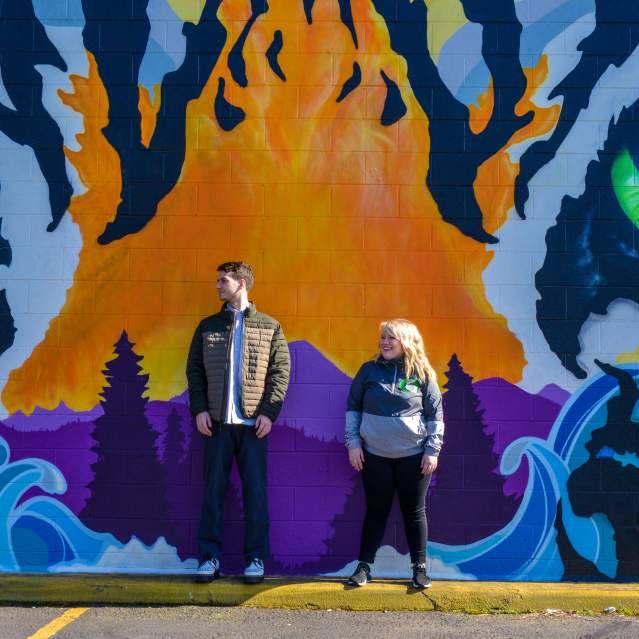 A man and woman stand in front of a brightly colored mural of a tiger's face.