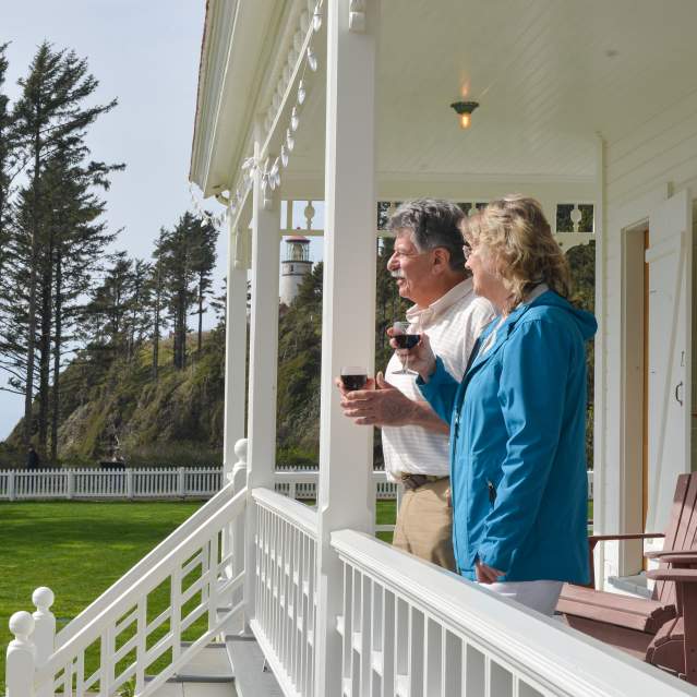 A man and a woman with wine glasses stand on a porch looking out at the ocean with a lighthouse in the distant background.