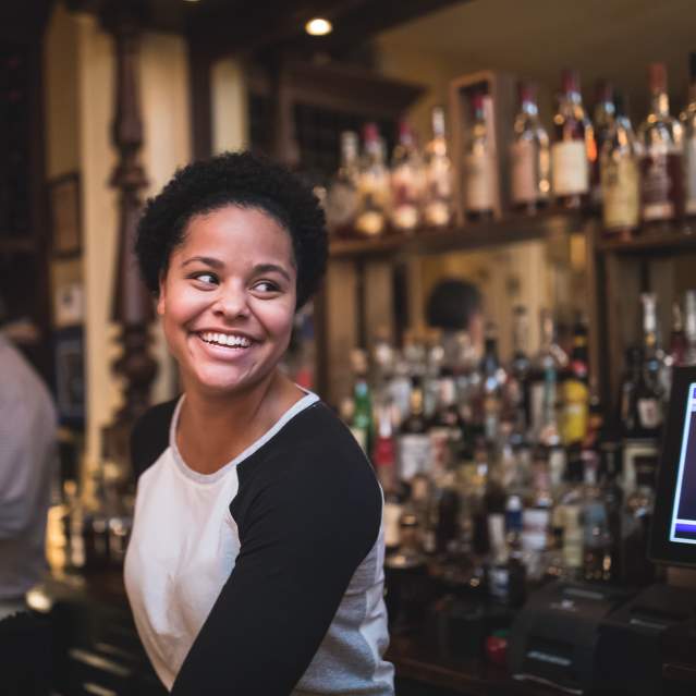 Young Black woman smiling with the bourbon collection of Tousey House Tavern behind her