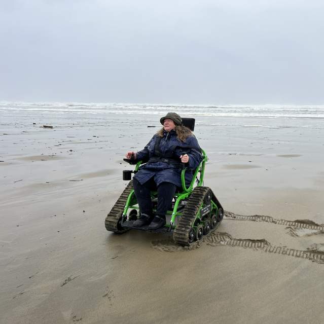 A person uses a green track chair on the flat, wet Oregon Coast beach.