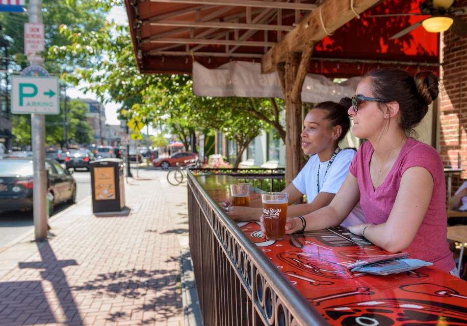 Two people sitting at a colorful outdoor bar table with drinks, beside a sidewalk on Main Street, Newark, Delaware, with cars and parking signs.