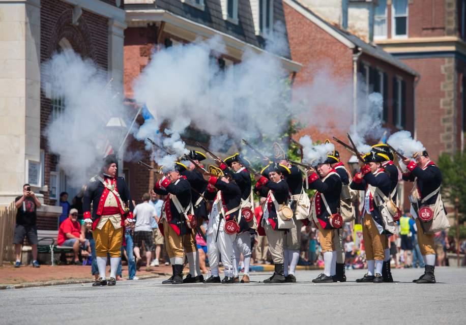 A group of historical reenactors in colonial military uniforms fires muskets, creating clouds of smoke during a lively outdoor event.