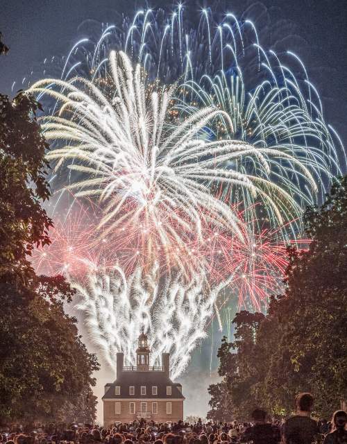 Fireworks illuminate the night sky above a historic building, surrounded by a crowd of spectators. Lush trees frame the scene, enhancing the festive atmosphere.