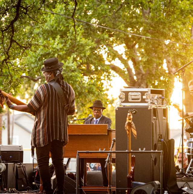 Booker T. Jones (on the piano) and his band members. The image is taken from the back of the stage and there is music equipment and speakers throughout the stage.