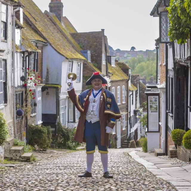 Rye cobbled street with Town Crier holding up his bell wearing traditional costume
