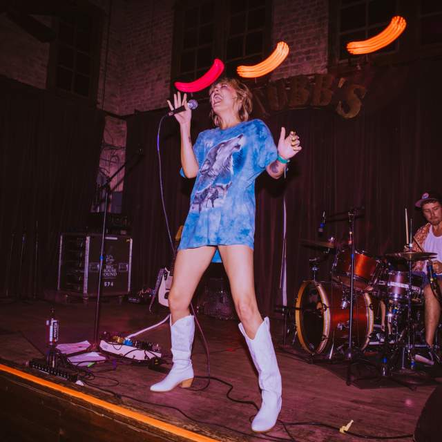 Woman wearing an oversized t-shirt as a dress with tall, white cowboyboots singing on the wooden stage at Stubb's.