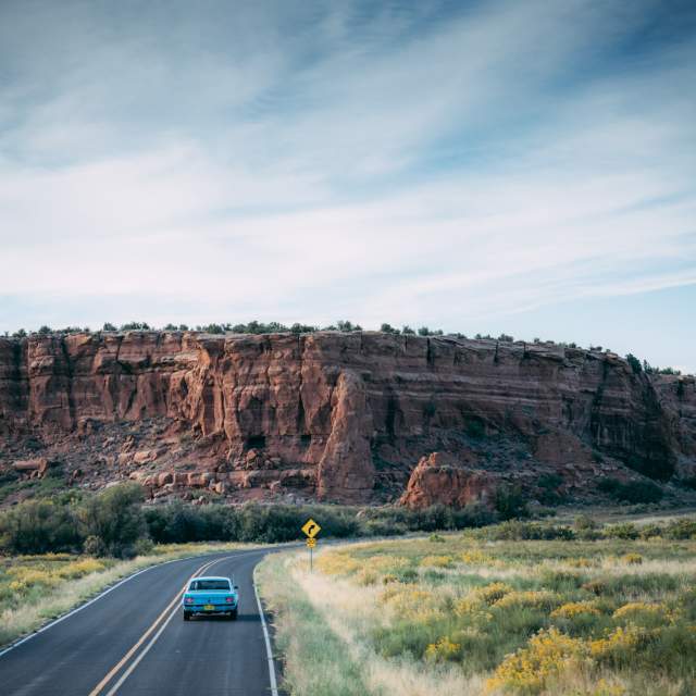 A car drives on a winding road past a large red rock formation under a clear blue sky.