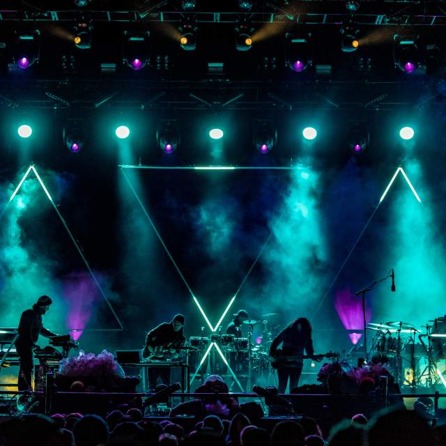Band performing on a dark, foggy stage lit with intense blue, purple, and yellow lights