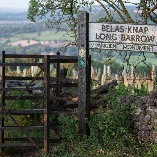 Kissing gate with footpath sign to Belas Knap with a stone wall ahead and behind a view of the valley