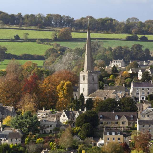 The town of Painswick in the Cotswolds, with it's church steeple rising in the centre, seen from a distance and surrounded by green rolling hills and trees