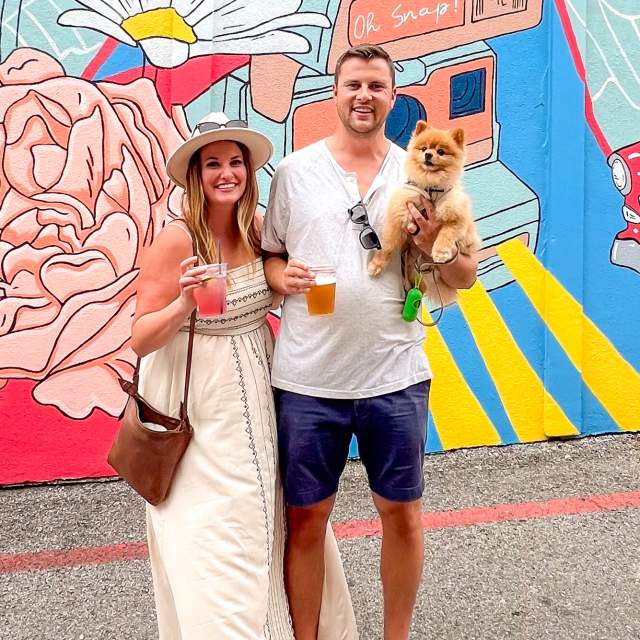 Couple standing in front of a colorful mural with cocktails and their little brown dog