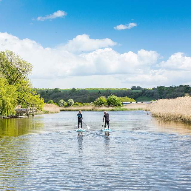 Two paddle boarders on the River Arun, with a boat moored to the left and grassy bank to the right, in Sussex