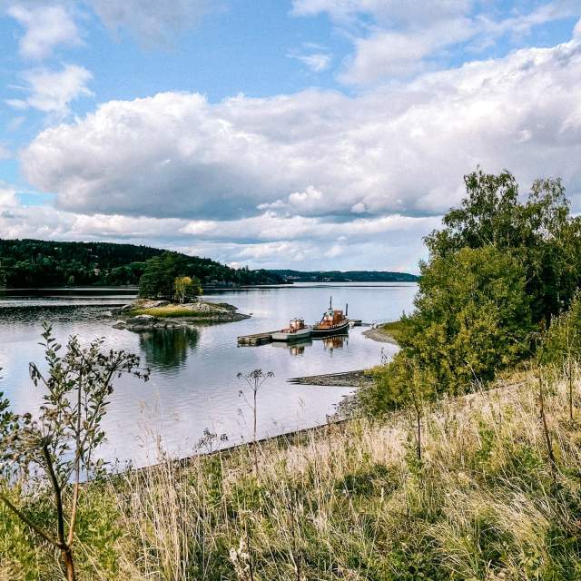 Utsikt over Glomma fra Furuholmen. To gamle båter ligger til kai langs ei flytebrygge.