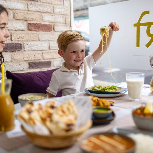 Child holding up noodles on a fork around table with family at Padharo