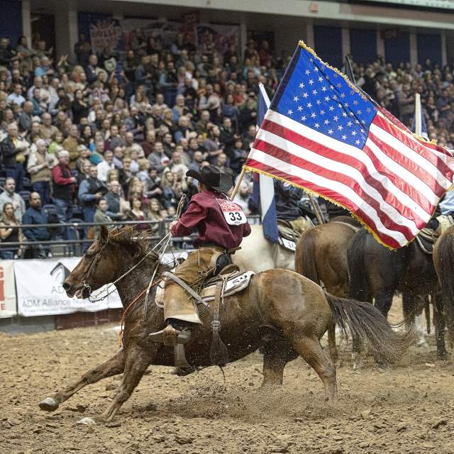 cowboys on horseback carry a large American flag across a packed Amarillo rodeo arena, dirt kicking up as the crowd watches from the stands