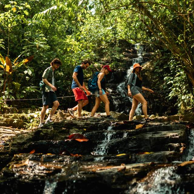 A group of hikers crossing a stream near Pozo Azul Waterfall, surrounded by many trees and nature.