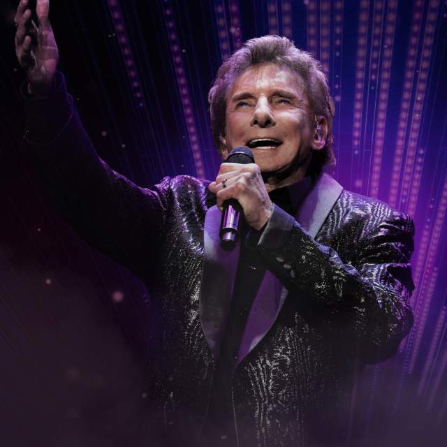 Barry Manilow singing into a microphone with his opposite hand extended out