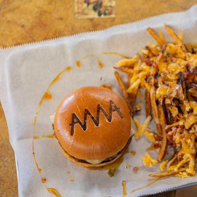 A juicy burger branded with “AMA” on the bun sits beside a hearty pile of fries loaded with melted cheese and bacon, served on a tray lined with parchment at a casual Amarillo eatery.